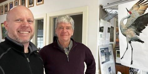 Selfie of two men smiling in a town hall with a painting of a crane in the background.